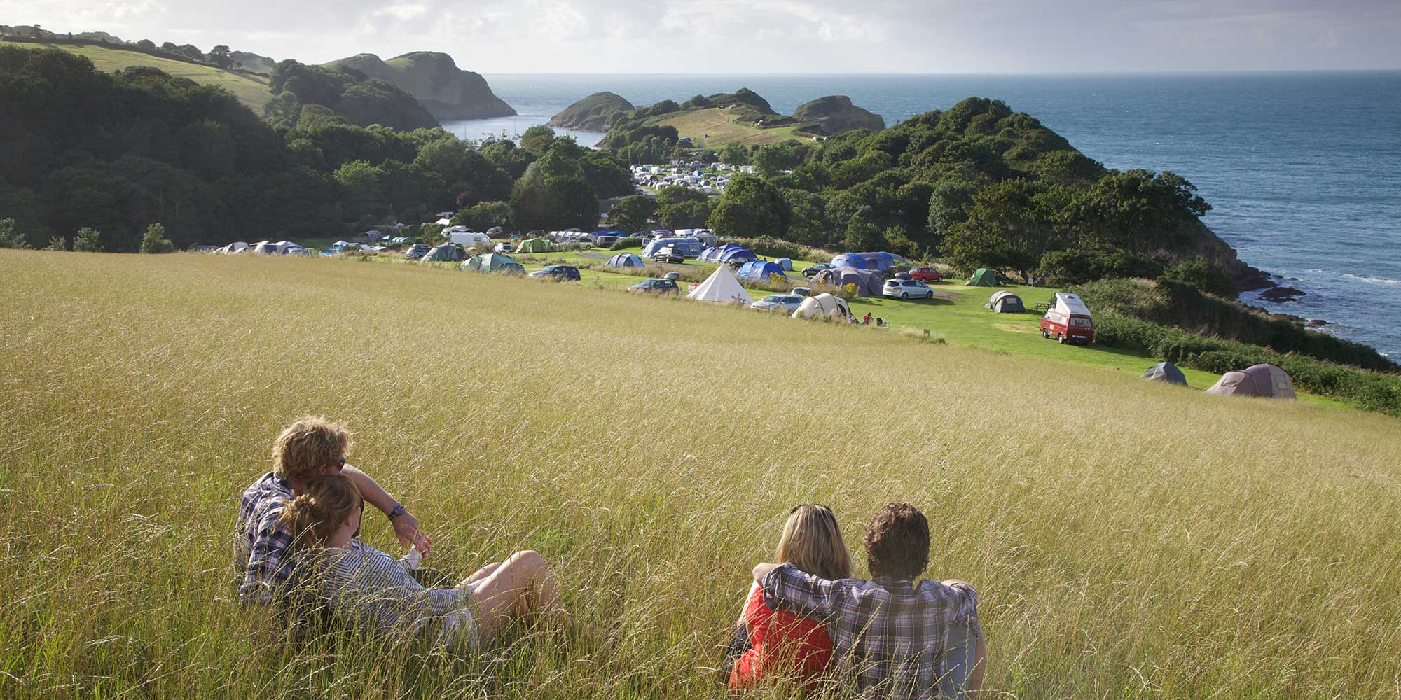 Two couples sitting in long grass looking down on a campsite next to a sea cliff