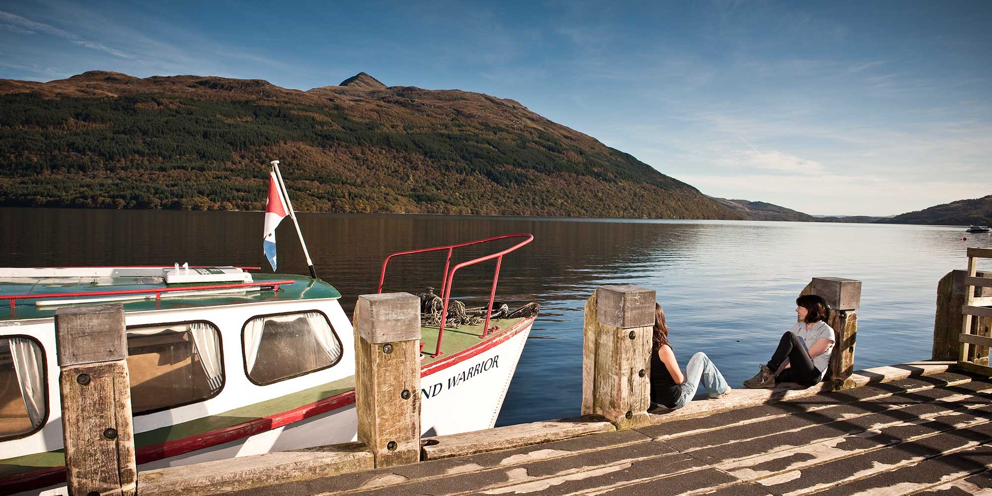 People sitting on a sun-drenched wooden jetty looking over a loch with mountains on the far shore